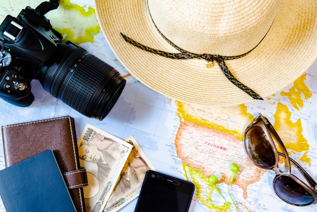 広げた世界地図の上に麦わら帽子・一眼レフカメラ・サングラス・パスポート・財布・紙幣・スマートホンを置いている