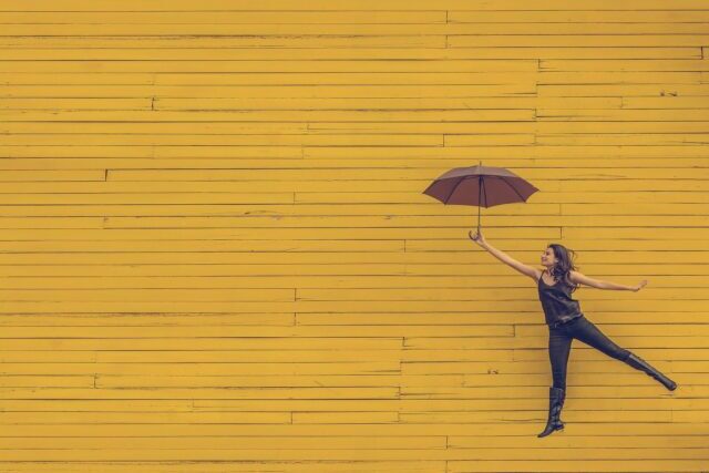 黄色い壁の前で傘を持って飛ぶ女性
